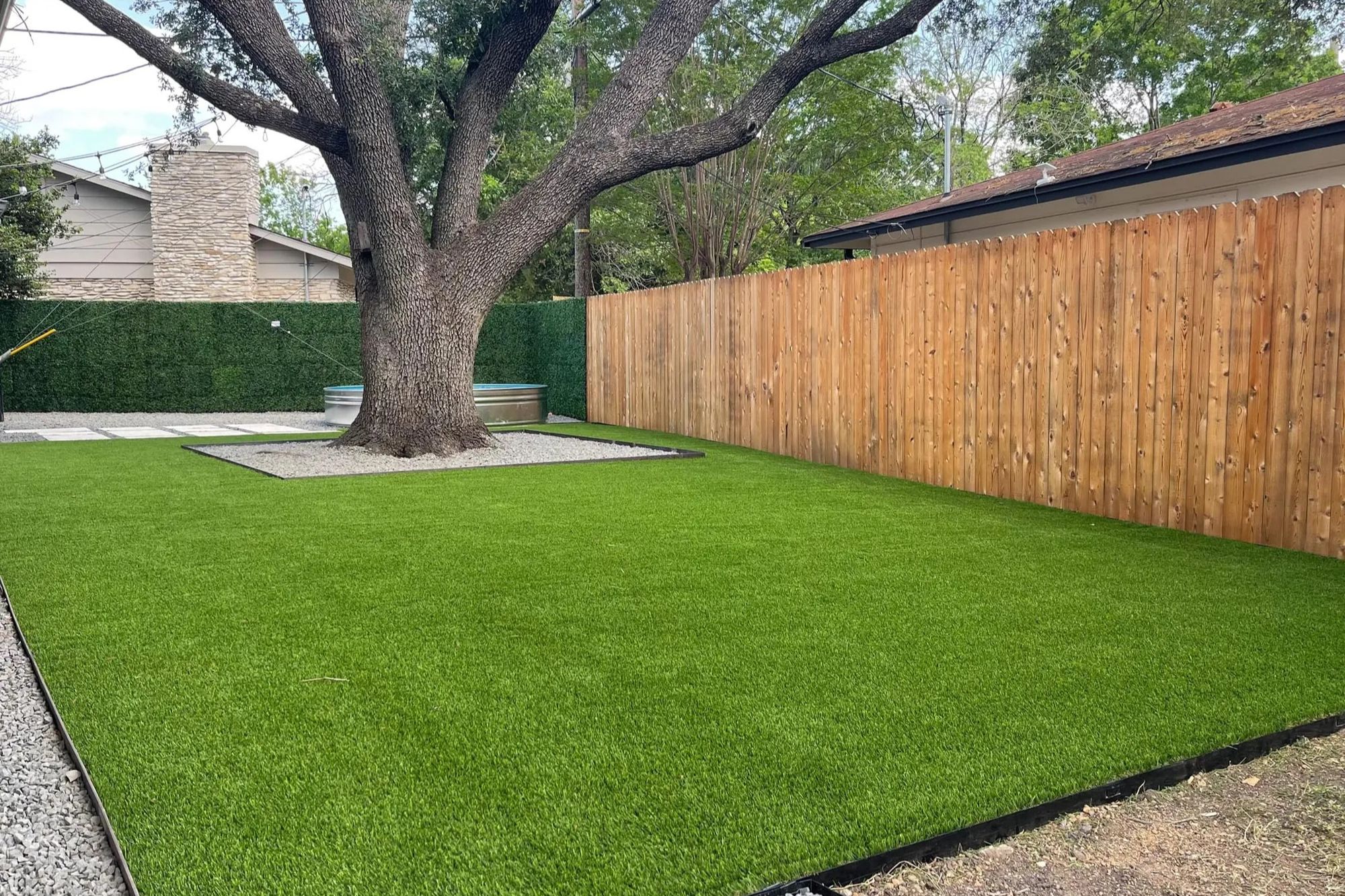 Modern backyard with artificial turf, gravel pathways, and a large central tree.